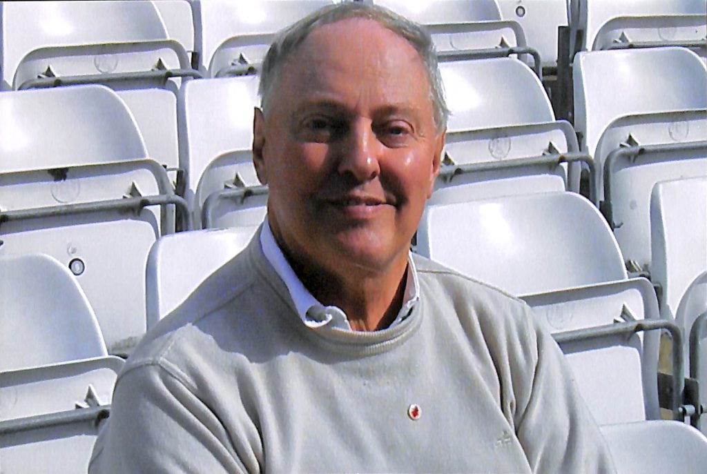 Robert Brooke smiling in sunshine; he is sitting in a stand, surrounded by upturned white seats.